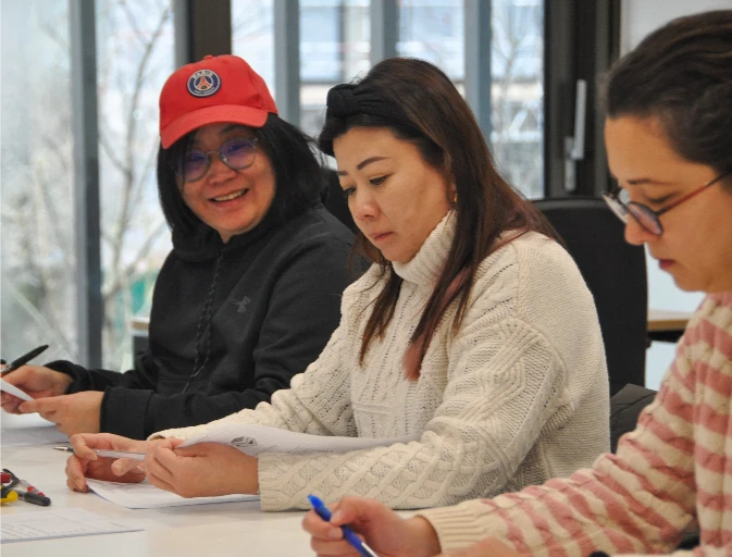 Three adult learners sitting together at a table, working on paperwork and smiling during a class