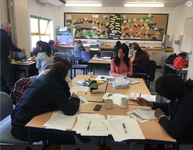 Adult learners working through English worksheets at tables in a bright classroom while their tutor supports them