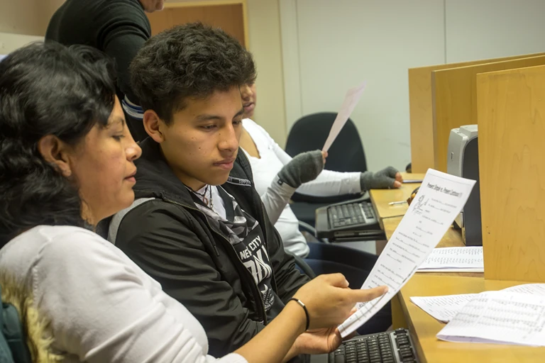 A young adult learner reading a course worksheet at a computer while a tutor supports him