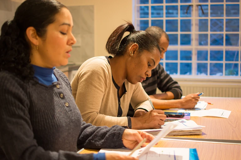 Three adult learners writing at desks in a classroom